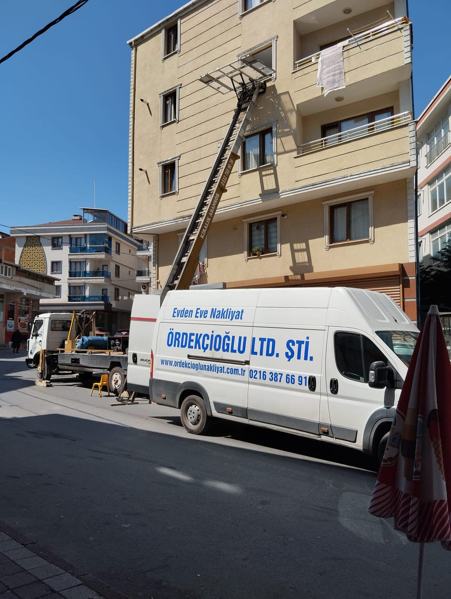 Apartman cephesine dayanmış dış cephe asansörü ve önünde “Evden Eve Nakliyat — Ördekçioğlu Ltd. Şti.” yazılı beyaz panelvan.