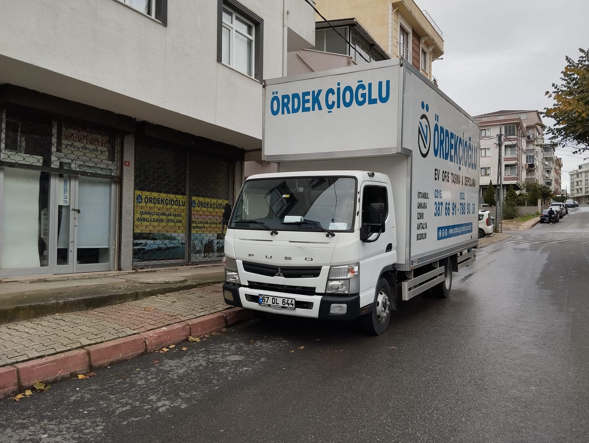 Üzerinde Ördekçioğlu yazısı ve telefon numaraları olan beyaz kapalı kasa kamyon, yağmur sonrası bir binanın önünde park hâlinde.