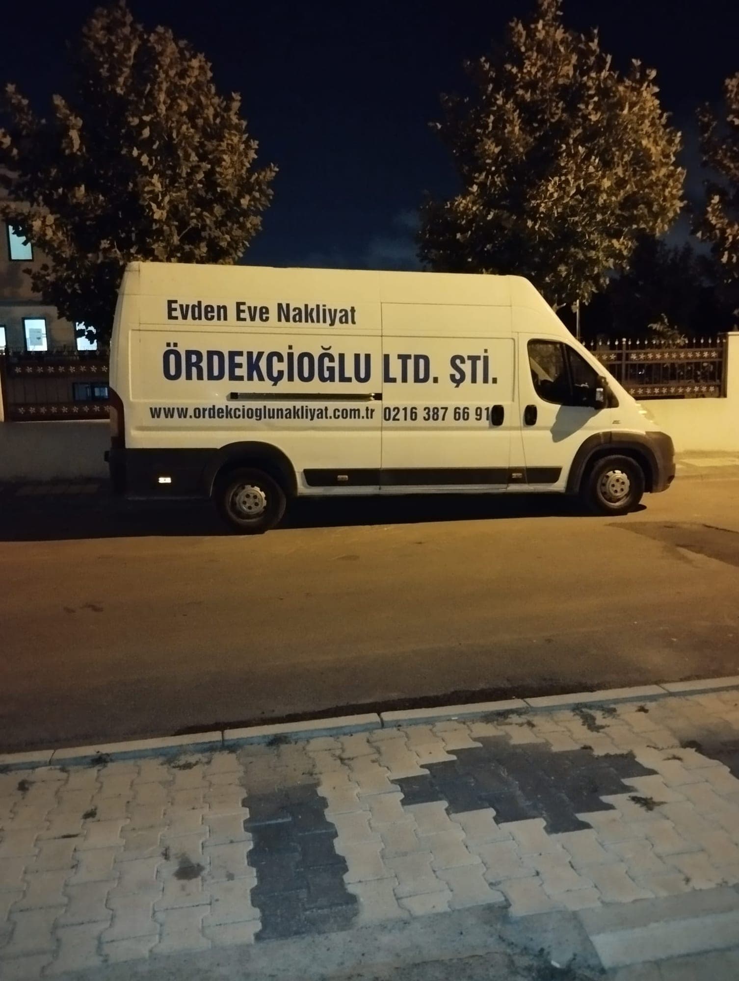 Gece çekimi: “Evden Eve Nakliyat — Ördekçioğlu Ltd. Şti.” yazılı beyaz panelvan, ağaçlı bir sokakta park hâlinde.