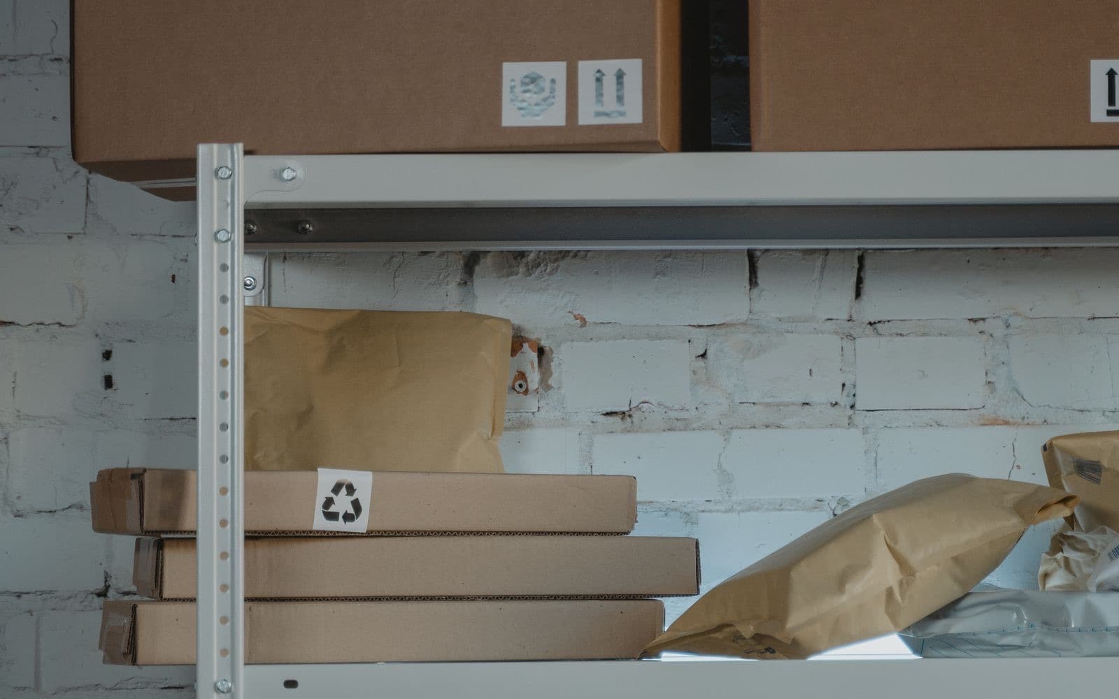 Beyaz tuğla duvar önündeki metal depo rafında taşıma işaretli karton koliler, düz karton kutular ve kraft kağıt paketler diziliyor.